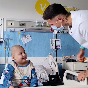 Un hombre con uniforme blanco y mascarilla mira a un niño con pijama azul sentado en una cama hospitalaria con equipos médicos al fondo