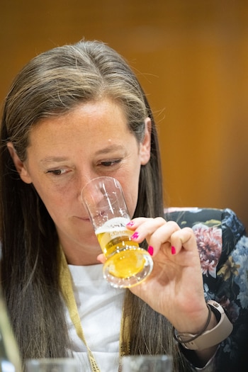 Mujer con cabello castaño y canas, camisa blanca y chaqueta floral, sujeta un vaso con cerveza clara para olerla. Usa un reloj y tiene las uñas rosadas