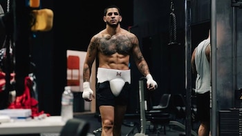Un hombre con tatuajes en el pecho, vendajes en las manos y protector pélvico camina con el torso descubierto en un gimnasio