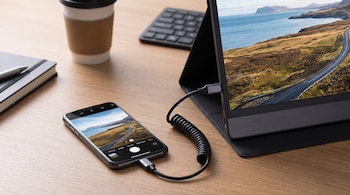 Teléfono móvil conectado por un cable negro en espiral a una pantalla externa que reproduce la misma fotografía sobre un escritorio.