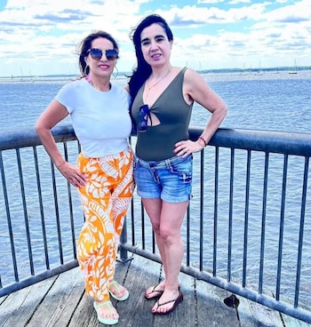 Dos mujeres de pie en un muelle de madera con barandilla metálica, con un cuerpo de agua y veleros al fondo bajo un cielo nublado