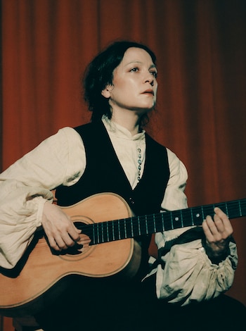 Natalia Lafourcade, de perfil, tocando una guitarra acústica con una camisa de mangas abullonadas y chaleco oscuro sobre un fondo rojo