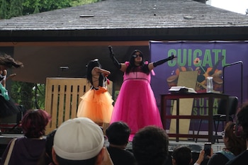 Dos performers en un escenario. Una lleva un vestido rosa vibrante, guantes negros, gafas de sol y micrófono. La otra, un top negro y tutu naranja