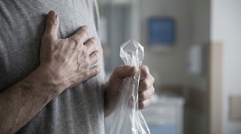 Dos manos de adulto con venas visibles, una sujeta una bolsa de plástico transparente, la otra sobre el pecho con camiseta gris. Fondo hospitalario.