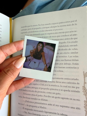Una mano sostiene una foto polaroid con la imagen de una mujer sonriente con pelo oscuro y remera negra, tomando mate, sobre un libro abierto