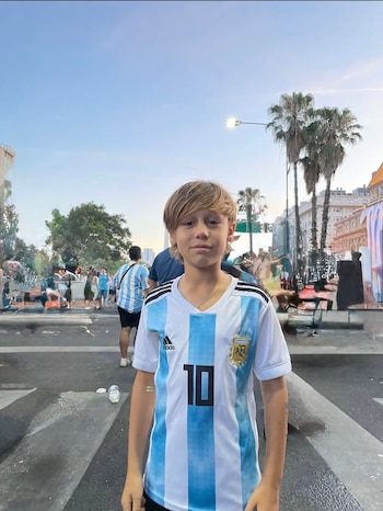 Un niño rubio con la camiseta blanca y celeste de Argentina con el número 10 en el pecho posa en una calle urbana. Al fondo se ven palmeras y personas
