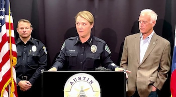 Mujer oficial de policía de uniforme azul oscuro habla en un podio con el logo de Austin; a su lado, un hombre de uniforme y otro de traje. Bandera de EE. UU. a la izquierda
