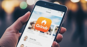 Una mano sostiene un smartphone con la aplicación Grindr abierta. La pantalla muestra el logo naranja de Grindr y perfiles de usuario borrosos al fondo.