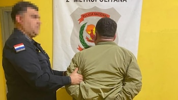 Agente de policía con uniforme y parche de la bandera de Paraguay en el brazo izquierdo, escoltando a un hombre de espalda con chaqueta caqui, frente a un cartel policial