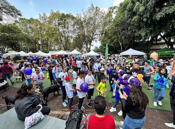 Una multitud de personas y perros en un espacio al aire libre con árboles, carpas blancas y globos morados