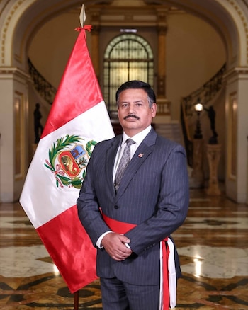 Hombre vestido con traje de raya diplomática y faja roja, de pie, delante de la bandera de Perú y un fondo con escaleras y arcos