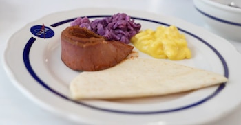 Plato blanco con borde azul y logo de NASA contiene un trozo de carne rojiza, repollo morado, puré amarillo y una tortilla doblada de color claro
