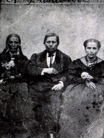 Fotografía antigua en blanco y negro de tres personas sentadas. Un hombre con barba y traje oscuro en el centro, flanqueado por dos mujeres
