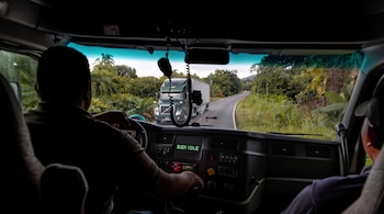 Interior de la cabina de un camión con dos personas frente al parabrisas, que muestra una carretera rural y un camión en el carril opuesto.