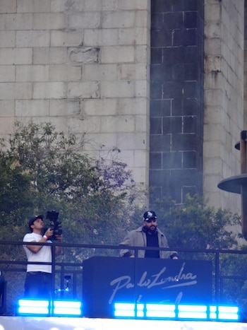Dos hombres sobre una tarima con luces azules y un cartel con el texto Paulo Londra, junto a árboles y una pared de bloques de piedra