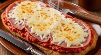 Primer plano de una milanesa a la napolitana con salsa de tomate, jamón, y queso mozzarella fundido, humeante en una fuente de cerámica con cubiertos a un lado.