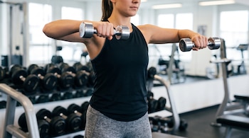 Una mujer con camiseta negra sostiene dos mancuernas metálicas a la altura del pecho frente a un soporte de pesas en un gimnasio.