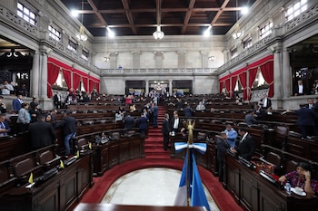 Un salón legislativo con curules de madera, alfombra roja, una bandera guatemalteca en primer plano y personas de traje