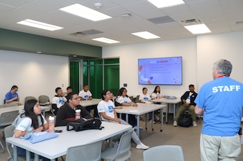 Un instructor con camiseta azul que dice "STAFF" se dirige a un grupo de jóvenes sentados en mesas en un aula. Una pantalla grande detrás muestra una presentación sobre "WHAT IS CRISPR?"
