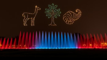Figuras luminosas de vicuña, árbol y cornucopia sobre fuentes de agua de colores rojo, azul y morado, de noche