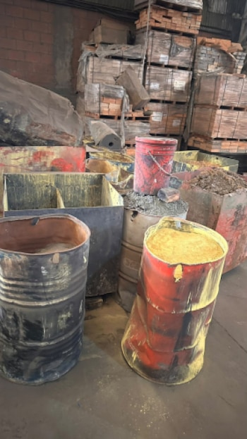 Tambores metálicos con polvo amarillo y oscuro frente a cajas metálicas y tarimas apiladas con bloques en un galpón