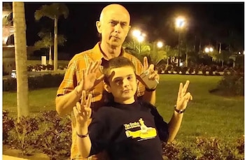 Hombre calvo con camisa a cuadros naranja y niño con camiseta negra de The Beatles posan haciendo el signo de la paz. Fondo nocturno con palmeras y luces