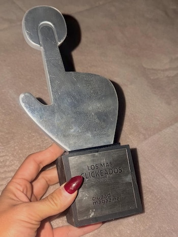 Una mano con uñas pintadas de rojo oscuro sostiene un trofeo plateado con forma de puntero de ratón, que dice "Los Más Clickeados 2023" y "ciudad magazine"