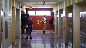 Vista de un pasillo escolar con estudiantes, paredes de ladrillo claro y una pared roja con el número 35 en amarillo; se ven lockers y un cartel de EXIT