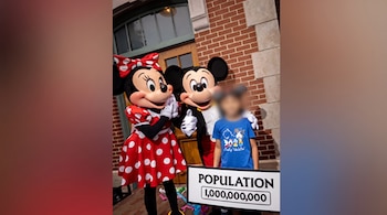 Minnie Mouse e Mickey Mouse junto com crianças com rosto confuso. Uma faixa branca diz 'POPULAÇÃO 1.000.000.000'. Base: parede de tijolos.