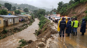 Río desbordado con agua turbia y orillas erosionadas, junto a una carretera con vehículos de emergencia y personal en chubasqueros; se ven casas en una ladera.
