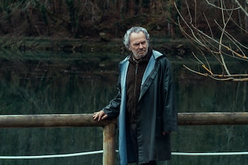 Un hombre con cabello gris, barba, abrigo oscuro y suéter marrón se apoya en una barandilla de madera frente a un lago con árboles