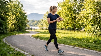 Mujer vestida con ropa deportiva oscura camina por un camino de asfalto en un parque mientras mira un reloj digital en su muñeca.