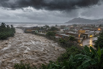 Vista panorámica de un río de agua marrón caudalosa bordeando casas bajas junto al mar bajo un cielo nublado y lluvia.