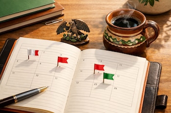 Agenda abierta en escritorio de madera con banderas pequeñas de colores rojo, verde y blanco, pluma, figura de águila, taza de café humeante y libros.