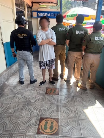 Hombre con rostro pixelado, oficial de INTERPOL SGIC con chaleco negro, tres oficiales de INM con uniformes verde olivo. Letrero de 'MIGRACION'. Piso de baldosas, marcadores de distancia