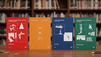 Cuatro libros de colores rojo, naranja, azul y verde colocados sobre una mesa de madera frente a una biblioteca desenfocada