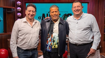 Tres hombres de pie y sonrientes posan juntos abrazados por los hombros en un estudio de televisión frente a una pantalla.