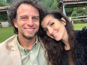 Hombre de pelo rizado y barba y mujer de pelo largo posan sonrientes. El fondo muestra vegetación y construcciones rústicas