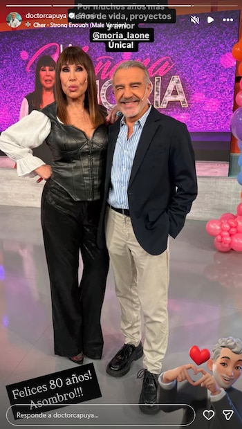 Moria Casán y Doctor Capuya posan de pie sonriendo. Moria Casán viste blusa blanca y conjunto de cuero negro. Doctor Capuya lleva blazer azul y camisa rayada