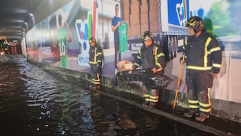 Tres bomberos con uniformes negros y franjas amarillas reflectantes se paran en un paso a desnivel con el suelo cubierto de agua oscura. Uno sostiene una pieza de vehículo. Un gran cartel publicitario se ve al fondo