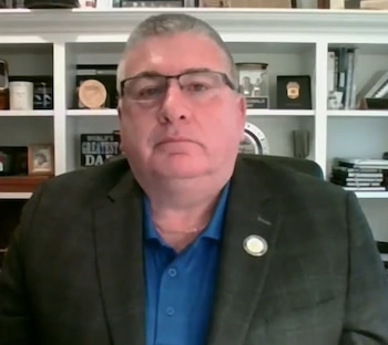 Un hombre de mediana edad, Bobby McDonald, con gafas y traje oscuro sobre camisa azul, sentado frente a una estantería blanca con libros y objetos