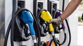 Un surtidor de combustible con tres mangueras de colores azul, amarillo y púrpura; una mano enguantada sujeta la manguera amarilla.