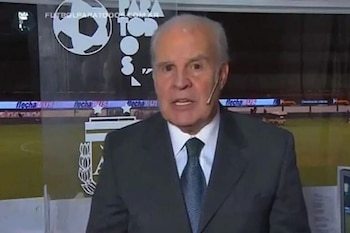 Primer plano del periodista Julio Ricardo con canas, vistiendo traje oscuro y corbata azul, con un micrófono de diadema, frente a un fondo con un balón de fútbol