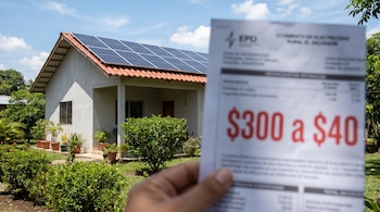 Primer plano de una mano sosteniendo una factura de electricidad con una reducción a $40, y una casa en El Salvador con paneles solares en el tejado bajo un cielo azul.