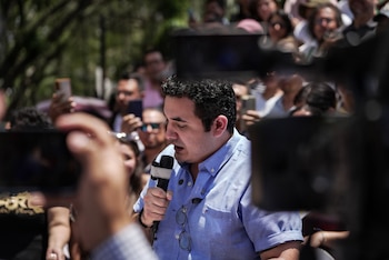 Primer plano de un hombre con camisa azul de manga corta hablando en un micrófono al aire libre, con gafas colgando de su cuello y una multitud borrosa detrás
