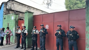 Una multitud reunida de noche en una calle frente a agentes de policía con escudos, con postes de luz y vehículos al fondo