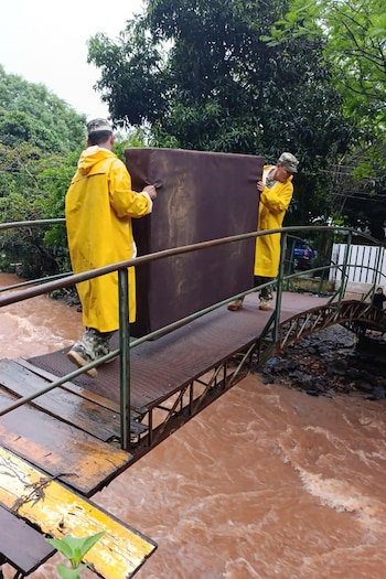 Dos hombres con impermeables amarillos cargan un colchón sobre un puente de metal situado sobre un río caudaloso de color marrón
