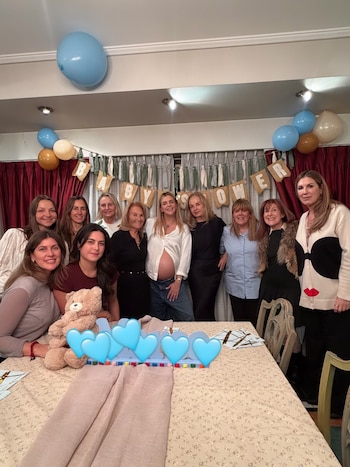Grupo de diez mujeres posando en una habitación con decoración de baby shower, incluyendo globos azules, dorados y blancos, y un cartel con letras