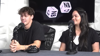 Un hombre y una mujer sentados frente a micrófonos en un estudio de podcast o radio, con una pantalla oscura al fondo que muestra gráficos en forma de dados