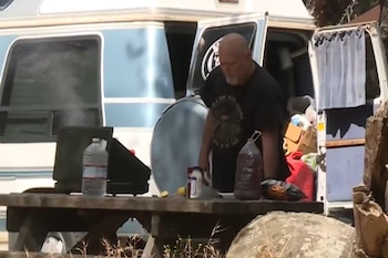 Un hombre con camiseta oscura permanece junto a una camioneta azul frente a una mesa de madera que tiene una cocina portátil encendida y botellas